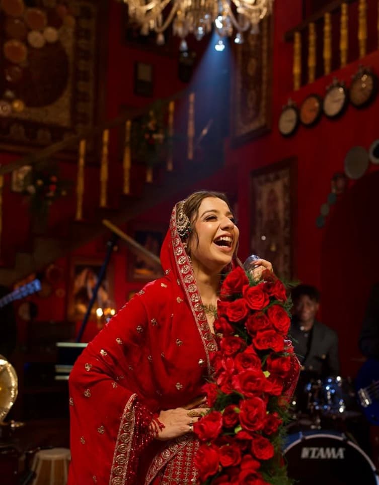 Jasmine Sandlas singing into a handheld mic in a red and gold outfit with a dupatta over her head, holding an armful of red roses, her band behind her